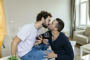 LGBTQ coppia Abbracciare una persona ogni altro e potabile vino al chiuso. Due romantico giovane maschio Gli amanti guardare a ogni altro mentre seduta insieme nel loro vivente camera. giovane gay coppia essere romantico a casa. foto