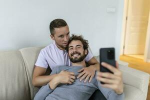 carino giovane gay coppia video chiamata loro amici nel loro vivente camera a casa. Due maschio Gli amanti sorridente allegramente mentre saluto loro amici su un' smartphone. giovane gay coppia seduta insieme. foto