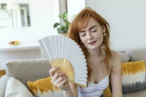 vicino su surriscaldato donna agitando carta fan, respirazione aria, pendente indietro su divano solo, sofferenza a partire dal il riscaldamento, febbre o caldo estate tempo metereologico a casa foto