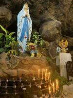 statua di santo vergine Maria nel romano cattolico Chiesa, nel il grotta di vergine Maria, nel un' roccia grotta cappella cattolico Chiesa con tropicale fiori in giro foto