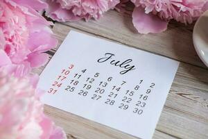 luglio calendario, peonie e un' tazza di caffè su il tavolo foto