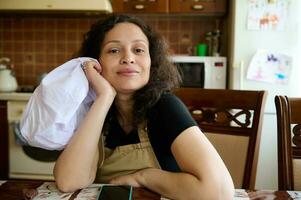 bellissimo giovane donna casalinga, panettiere pasticciere nel dello chef grembiule, seduta a tavolo nel casa cucina, sorridente a telecamera foto