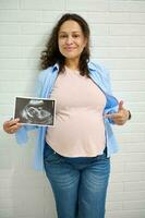 maturo contento incinta donna puntamento a sua gonfiarsi, Tenere il ecografia di sua bambino, sorridente guardare a telecamera foto