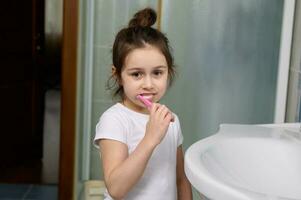 bellissimo bambino in età prescolare poco ragazza sorridente a telecamera mentre spazzolatura denti, in piedi di Lavello nel il casa bagno. foto