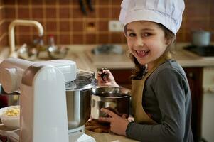 adorabile malizioso poco bambino ragazza degustazione fuso belga cioccolato, in piedi di cucina controsoffitto, sorrisi a telecamera foto