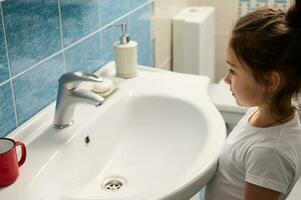 lato Visualizza poco bambino ragazza nel bianca pigiama, in piedi di Lavello nel il bagno durante quotidiano mattina igiene routine foto