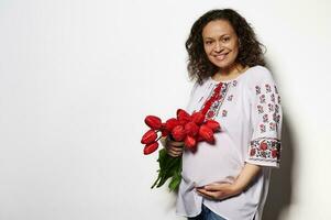 autentico incinta donna toccante sua gonfiarsi, sorridente guardare a telecamera, Tenere rosso tulipani, isolato su bianca sfondo foto