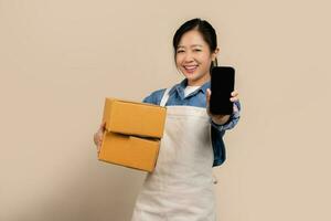 imprenditore asiatico donna indossare grembiuli su leggero Marrone sfondo. lei Tenere cartone scatole e mostrando vuoto schermo smartphone. foto