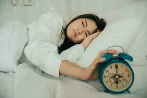 asiatico bellissimo donna nel bianca pigiama svolta via allarme orologio mentre assonnato nel letto a sua Camera da letto di Casa o Hotel su vacanze. ragazza è avendo guaio scia su in ritardo nel mattina. malsano dormire. foto