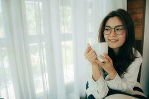 bellissimo giovane asiatico donna seduta di il finestra. lei è sorridente - Tenere un' tazza di caffè nel mano Diviso su nel ordine per inalare il aroma di caffè. mattina sole brilla attraverso il bicchiere finestra. foto