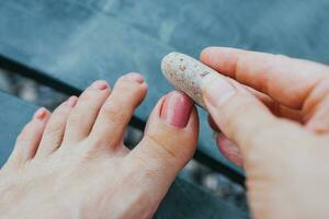 il ragazza su il spiaggia leviga e lucidi sua unghia del piede con un abrasivo pietra di ciottoli - indipendente pedicure a il mare foto