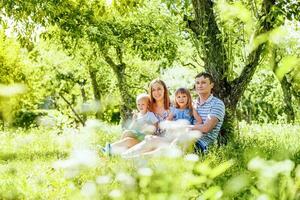 un' caucasico coppia seduta sotto un' albero con loro giovane figlio e un' figlia nel loro giri nel un' parco foto