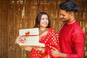 indiano uomo nel festivo etnico Abiti dando un' avvolto regalo scatola per un' donna nel un' rosso saree, lei è sorridente e sensazione contento foto