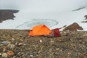panoramico alpino paesaggio con arancia rinforzata tenda e escursioni a piedi zaino con il trekking poli contro il sfondo di un' ghiacciaio su un' alta altitudine altopiano. viaggio concetto. foto