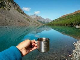 mano Tenere un' escursionisti tazza con pulito acqua. pulito potabile acqua nel il blu montagna lago. pulito acqua nel un' turista tazza contro il sfondo di un' lago e nevoso montagne. foto