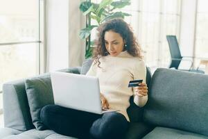 donna latina che utilizza laptop e mano che tiene la carta di credito per lo shopping sul divano foto