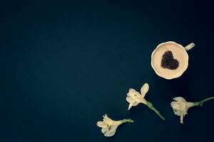 minimalista piatto posizione. tre fiori e caffè con crema e cannella nel il modulo di un' cuore nel un' boccale su un' buio blu sfondo. bianca con giallo petali. vignettatura. copia spazio su il sinistra. foto