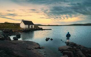 mozzafiato in riva al lago scenario con uomo in piedi su il roccia Guardando Alba sopra del pescatore capanna a screebe, connemara nazionale parco nel contea Galway, Irlanda foto