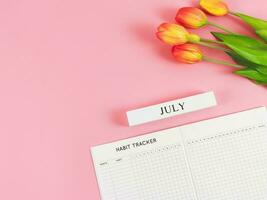 piatto posare di abitudine inseguitore libro con di legno calendario luglio, e tulipani su rosa sfondo. foto