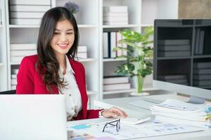 asiatico donna d'affari nel rosso completo da uomo calcolatore azienda i saldi con calcolatrice, computer portatile, e tavoletta su tavolo interno di donna d'affari ufficio a il moderno postazione di lavoro. foto