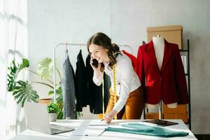 moda progettista donna parlando inteligente Telefono e utilizzando il computer portatile con digitale tavoletta computer nel moderno studio il Abiti sospeso su il cremagliere foto