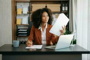 fiducioso bellissimo africano donna d'affari digitando il computer portatile computer e digitale tavoletta mentre Tenere caffè a ufficio foto