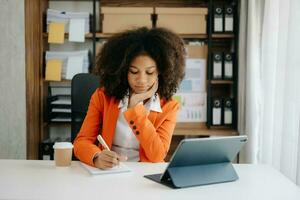 giovane africano donna digitando su tavoletta e il computer portatile mentre seduta a il Lavorando di legno tavolo moderno ufficio foto