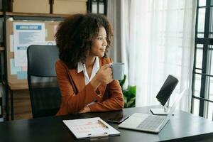 fiducioso bellissimo africano donna d'affari digitando il computer portatile computer e digitale tavoletta mentre Tenere caffè a ufficio foto