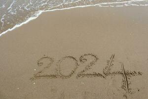nuovo anno 2024 scritto nel il sabbia su un' spiaggia con mare onda sfondo. foto