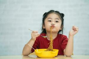 asiatico bambino ragazza godere contento utilizzando posate cucchiaio e forchetta mangiare delizioso spaghetto nel cucina su cenare tavolo. contento asiatico bambino ragazza pratica mangiare di sua se stesso su cenare tavolo. bambino cibo concetto foto