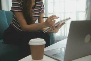 contento asiatico adolescente ragazza Tenere tampone computer aggeggio utilizzando digitale tavoletta tecnologia seduta su il divano a casa. sorridente giovane donna utilizzando app, shopping in linea, lettura notizia, navigazione Internet su divano foto