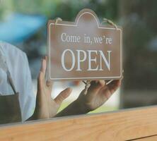 aprire. barista, cameriera donna indossare protezione viso maschera svolta Aperto cartello tavola su bicchiere porta nel moderno bar caffè negozio, bar ristorante, Al dettaglio negozio, piccolo attività commerciale proprietario, cibo e bevanda concetto foto