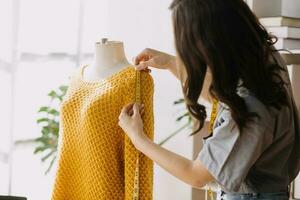 moda progettista l giovane asiatico donna Lavorando utilizzando computer portatile, tavoletta e sorridente mentre in piedi nel laboratorio rispondere su attività commerciale foto