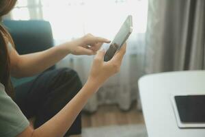 contento asiatico adolescente ragazza Tenere tampone computer aggeggio utilizzando digitale tavoletta tecnologia seduta su il divano a casa. sorridente giovane donna utilizzando app, shopping in linea, lettura notizia, navigazione Internet su divano foto