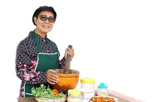 contento asiatico anziano donna è cucinando, indossare capocuoco berretto e grembiule, detiene pestello, mortaio e piatto di peperoncini. concetto, cucinando per famiglia. tailandese cucina stile di vita. anziano attività. foto