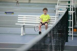 triste ragazzo si siede su un' panchina a un' tennis concorrenza. il bambino giochi tennis. foto