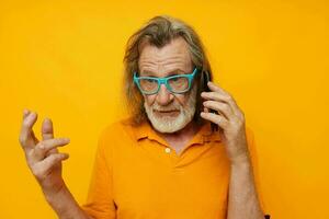 anziano dai capelli grigi uomo indossare blu bicchieri giallo camicia parlando su il Telefono isolato sfondo foto