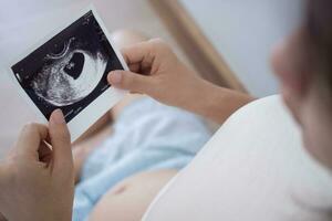 incinta donna è guardare a un ultrasuono foto di feto. madre delicatamente tocchi il bambino su stomaco. felice, famiglia, crescita, gravidanza, godimento , preparare neonato, prendere cura, assistenza sanitaria, pancia.