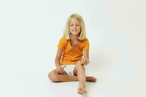 un' allegro poco ragazza nel un' giallo maglietta si siede su il pavimento con sua gambe piegato foto