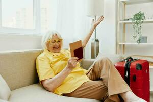contento anziano donna con passaporto e viaggio biglietto confezionato un' rosso valigia, vacanza e Salute cura. sorridente vecchio donna felicemente si siede su il divano prima il viaggio. foto
