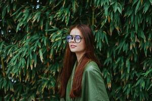 un' donna nel un' elegante di moda verde impermeabile e occhiali da sole passeggiate giù il strada vicino un' parco con bambù foto