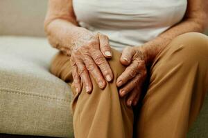 anziano donna acuto dolore nel sua gamba seduta su il divano, Salute i problemi nel vecchio età, povero qualità di vita. nonna con grigio capelli detiene su per sua dolorante ginocchio, i problemi con articolazioni e legamenti. foto