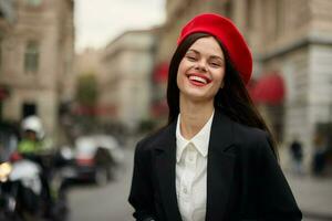 moda donna ritratto Sorridi denti in piedi su il strada nel il città sfondo nel elegante Abiti con rosso labbra e rosso berretto, viaggiare, cinematico colore, retrò Vintage ▾ stile, urbano moda stile di vita. foto