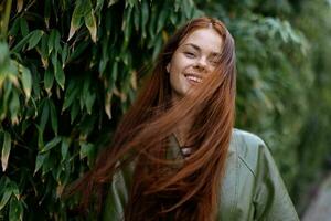 ritratto di felicità donna con rosso volante capelli Sorridi con denti a piedi nel il città nel il parco contro un' fondale di verde bambù foglie, primavera nel il città foto