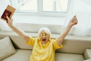 contento anziano donna con passaporto e viaggio biglietto confezionato un' rosso valigia, vacanza e Salute cura. sorridente vecchio donna con gioia seduta su il divano prima il viaggio sollevato sua mani su nel la gioia. foto