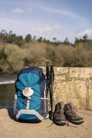 zaino con conchiglia, simbolo del camino de santiago con scarponcini da trekking e bastoncini appoggiati al muro di pietra foto