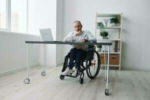 un' uomo nel un' sedia a rotelle, un' uomo d'affari nel il ufficio lavori a un' computer portatile, scrittura giù un' Piano nel un' taccuino, premurosità, integrazione in società, il concetto di Lavorando un' persona con disabilità foto