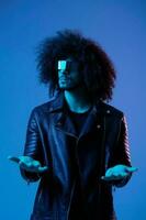 ritratto di moda uomo con Riccio capelli con elegante bicchieri su blu sfondo multinazionale, colorato luce, nero pelle giacca tendenza, moderno concetto. foto