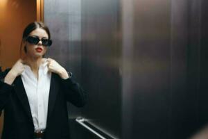 elegante donna nel un' nero giacca e occhiali da sole in posa nel un ascensore sollevamento vicino il specchio, moda modello, buio cinematico leggero e colore matrice, fascino Vintage ▾ foto