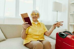 contento anziano donna con passaporto e viaggio biglietto confezionato un' rosso valigia, vacanza e Salute cura. sorridente vecchio donna con gioia seduta su il divano prima il viaggio sollevato sua mani su nel la gioia. foto